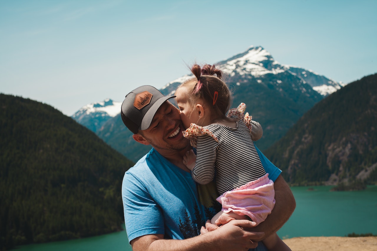 Man in blue shirt and black cap holding and smiling at a young girl who is kissing his cheek with a mountain and lake backdrop.