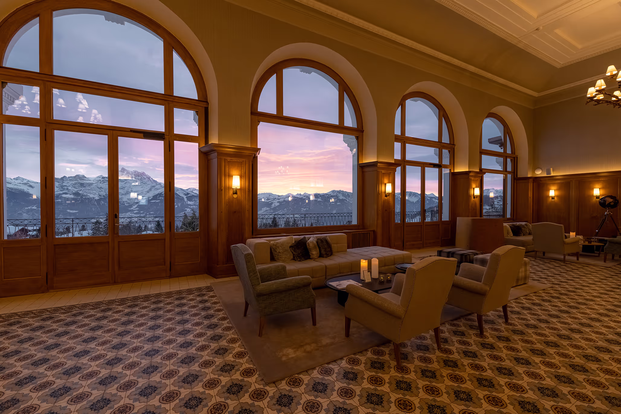 Elegant lounge area with comfortable seating and large arched windows showcasing a sunset view of snow-capped mountains.