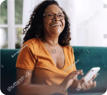 Smiling woman with curly hair and glasses using a smartphone on a green couch indoors.