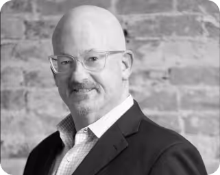 Black and white headshot of a bald man with glasses in a suit smiling slightly.