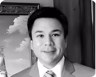 Smiling man in a suit and tie standing indoors with a framed picture and curtain behind him.