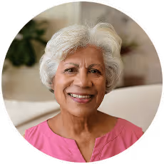 Smiling elderly woman with white hair wearing a pink top, sitting indoors with plants in the background.