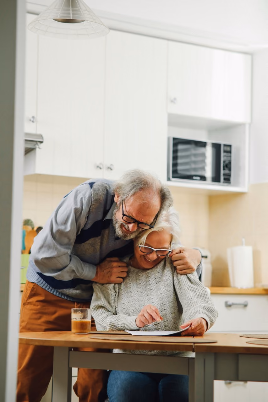 Elderly couple smiling and reviewing a document together at a kitchen table with coffee nearby.