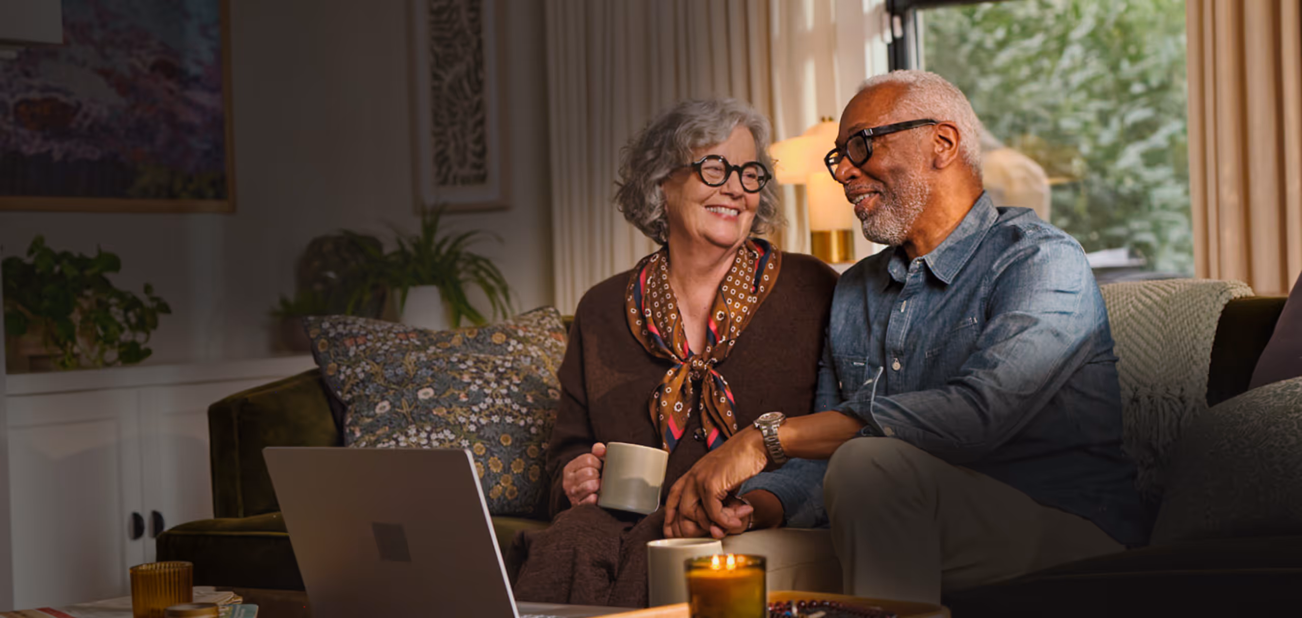 Senior couple sitting on a sofa, holding hands, smiling and looking at a laptop in a cozy living room.
