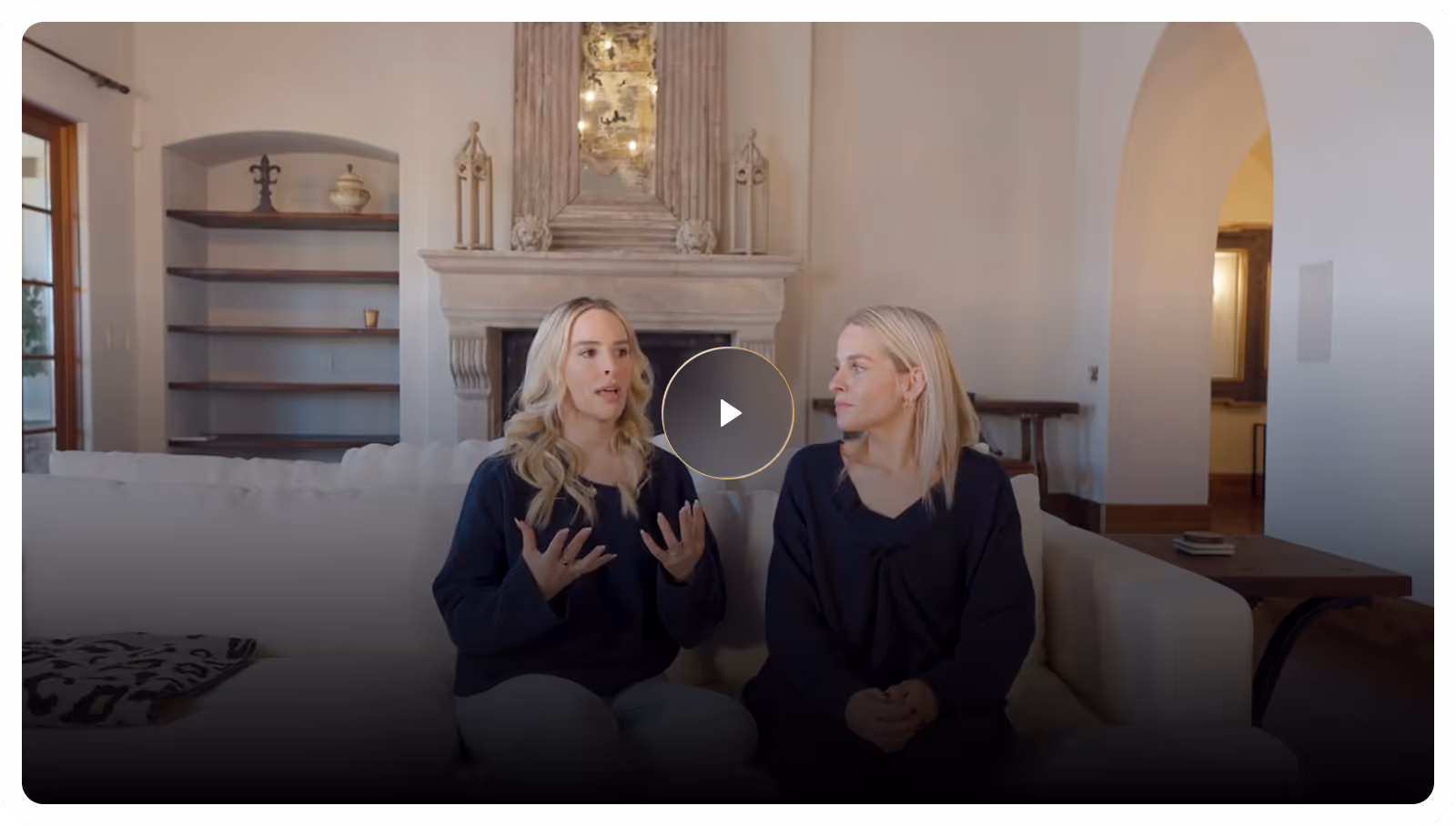 Two women sitting on a couch in a living room, one speaking and gesturing with hands.