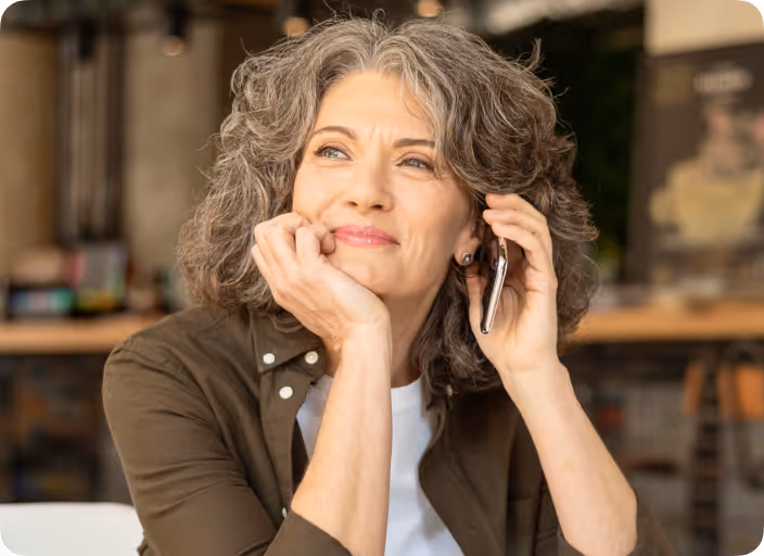 Smiling mature woman with gray hair holding a smartphone to her ear inside a cafe.