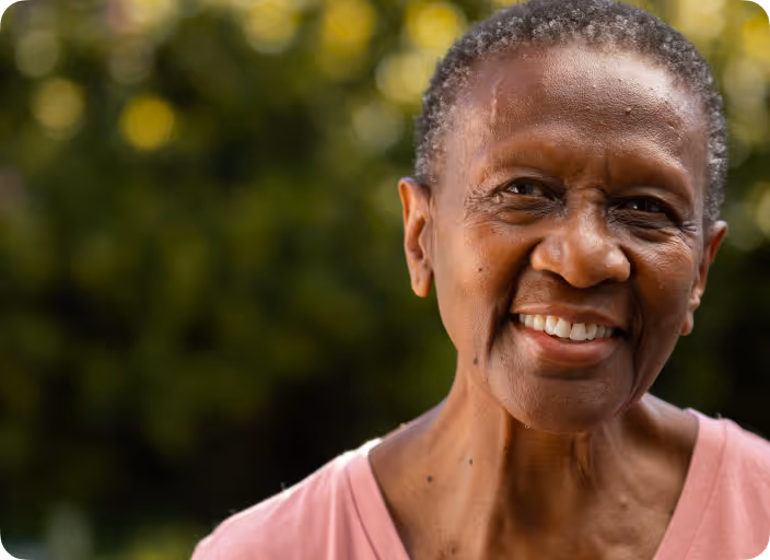 Smiling elderly woman with short hair wearing a pink shirt outdoors with blurred green background.
