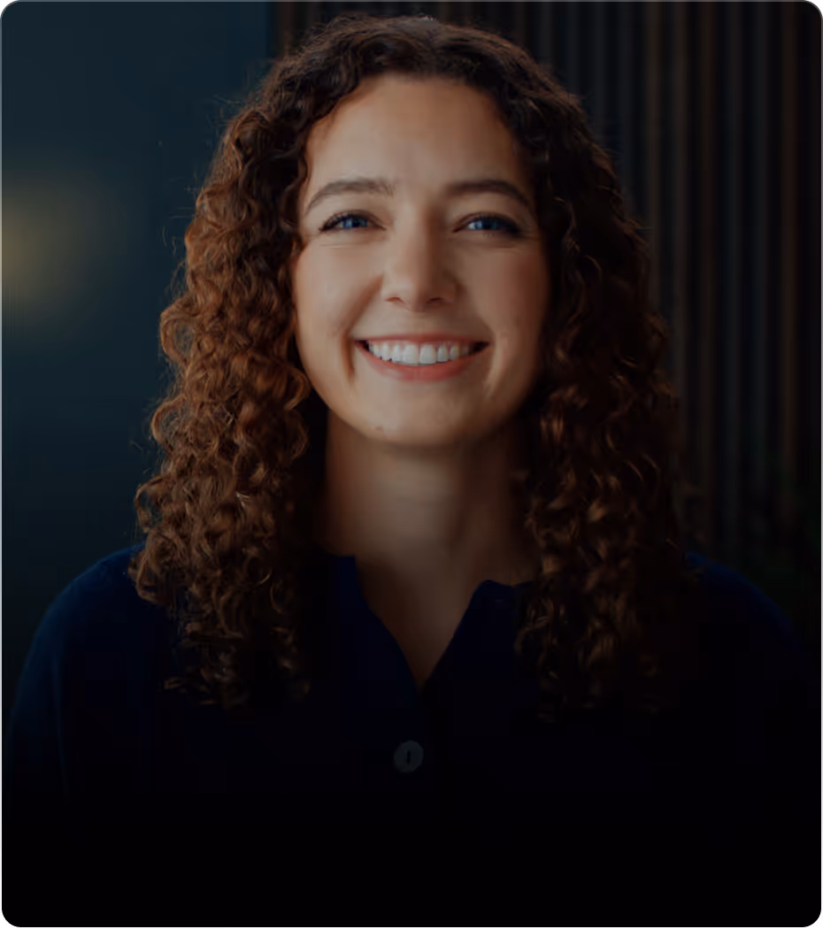 Smiling woman with curly hair and blue eyes wearing a dark top in a dimly lit room.
