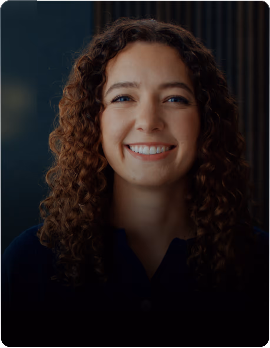 Smiling woman with curly hair wearing a dark top against a dark blurred background.
