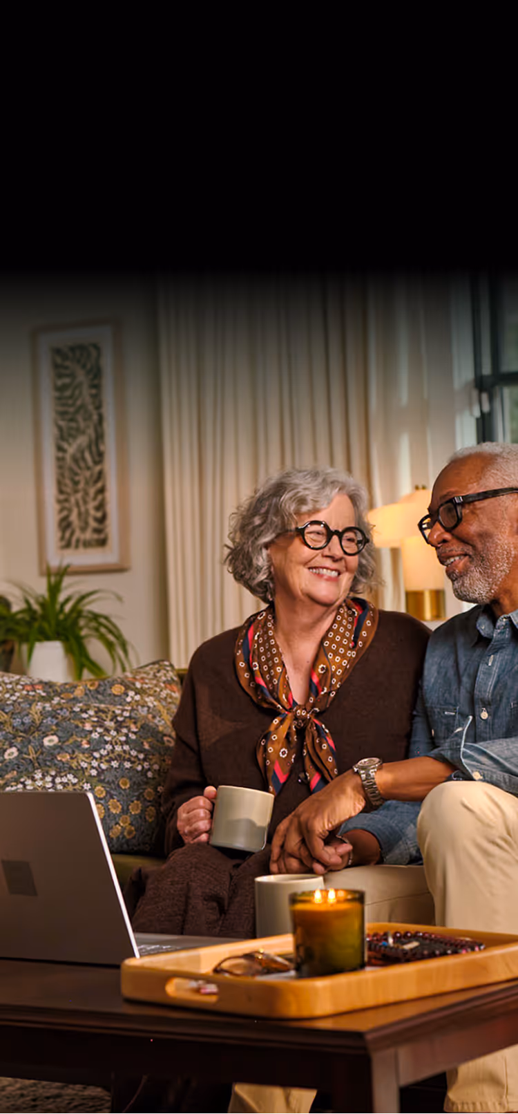 Elderly couple holding hands and smiling while sitting on a couch with coffee mugs and laptop nearby.