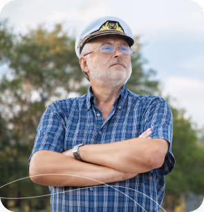 Older man in a captain's hat and blue plaid shirt with arms crossed outdoors.