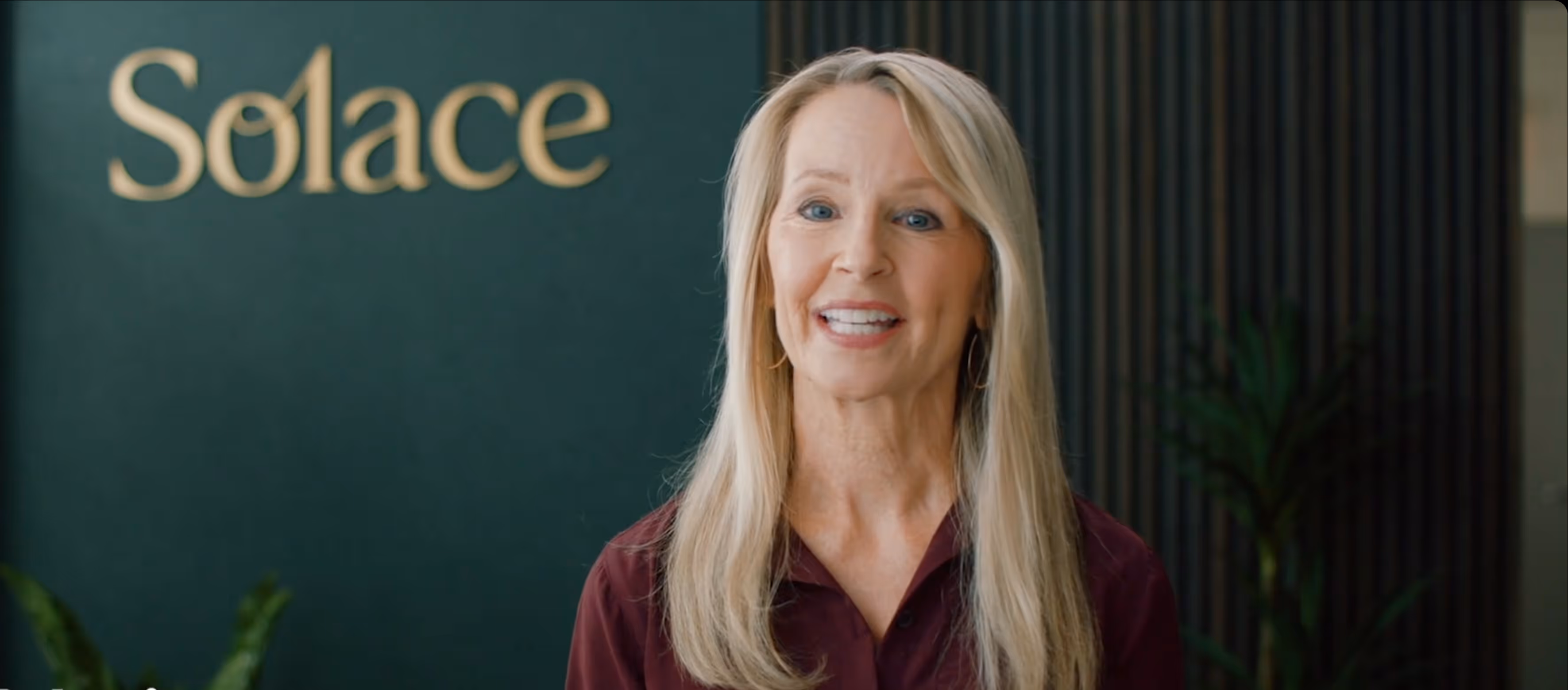 Smiling middle-aged woman with long blonde hair standing in front of a green wall with the word Solace.