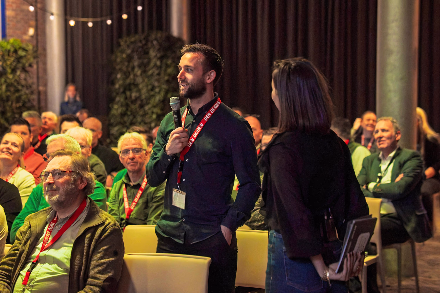 Man met microfoon spreekt tijdens bijeenkomst, vrouw en publiek luisteren in zaal.