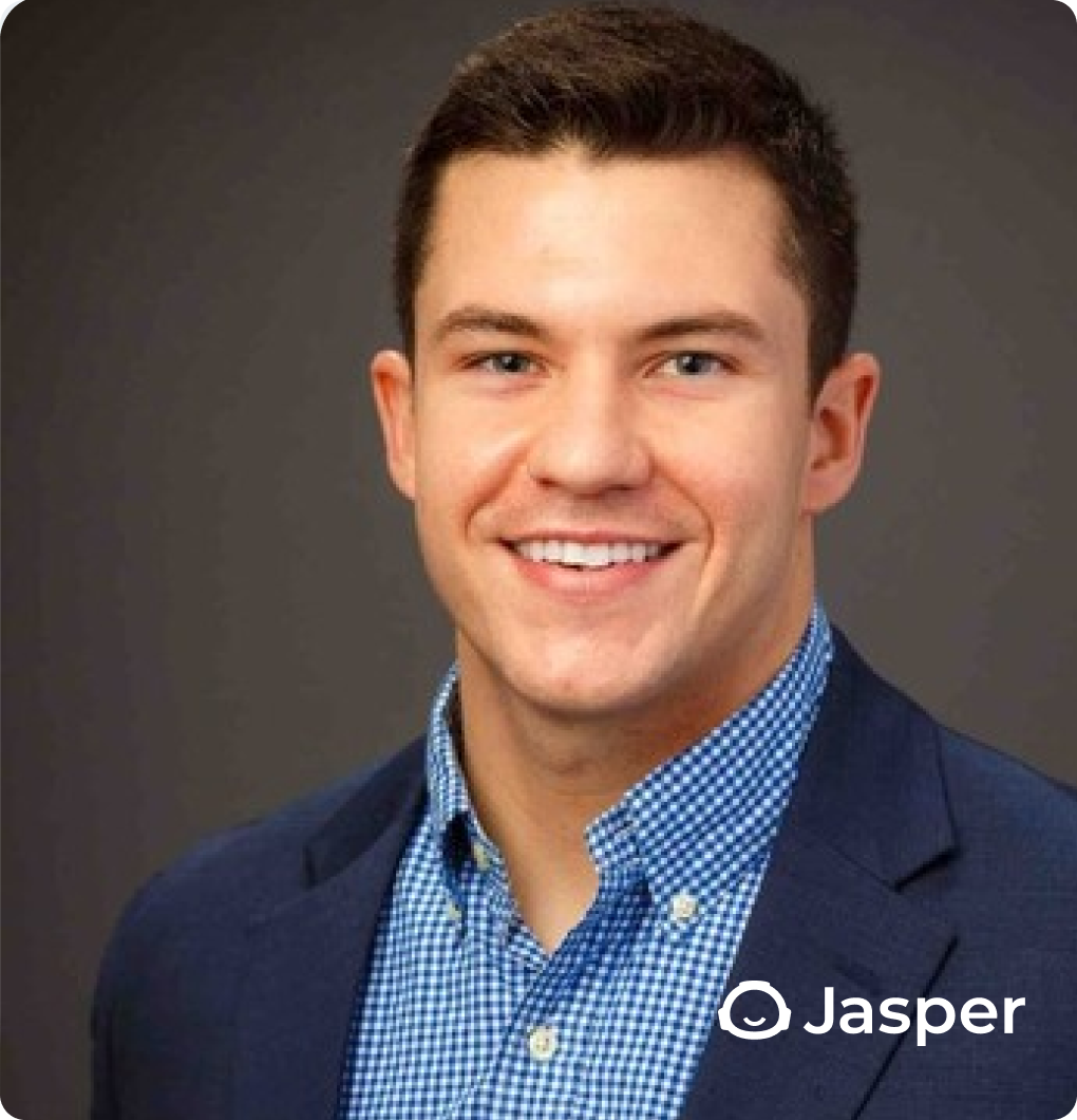 Smiling man in a navy blazer and blue checkered shirt against a gray background.
