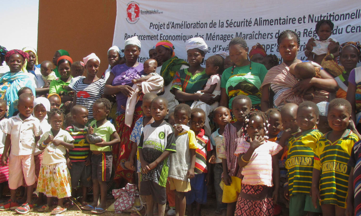 Eine große Gruppe von Müttern und Kindern in Burkina Faso steht vor einem Banner eines Ernährungsprojekts. Das Bild zeigt die breite Beteiligung an einem Programm zur Verbesserung der Nahrungssituation für Kinder in Burkina Faso.