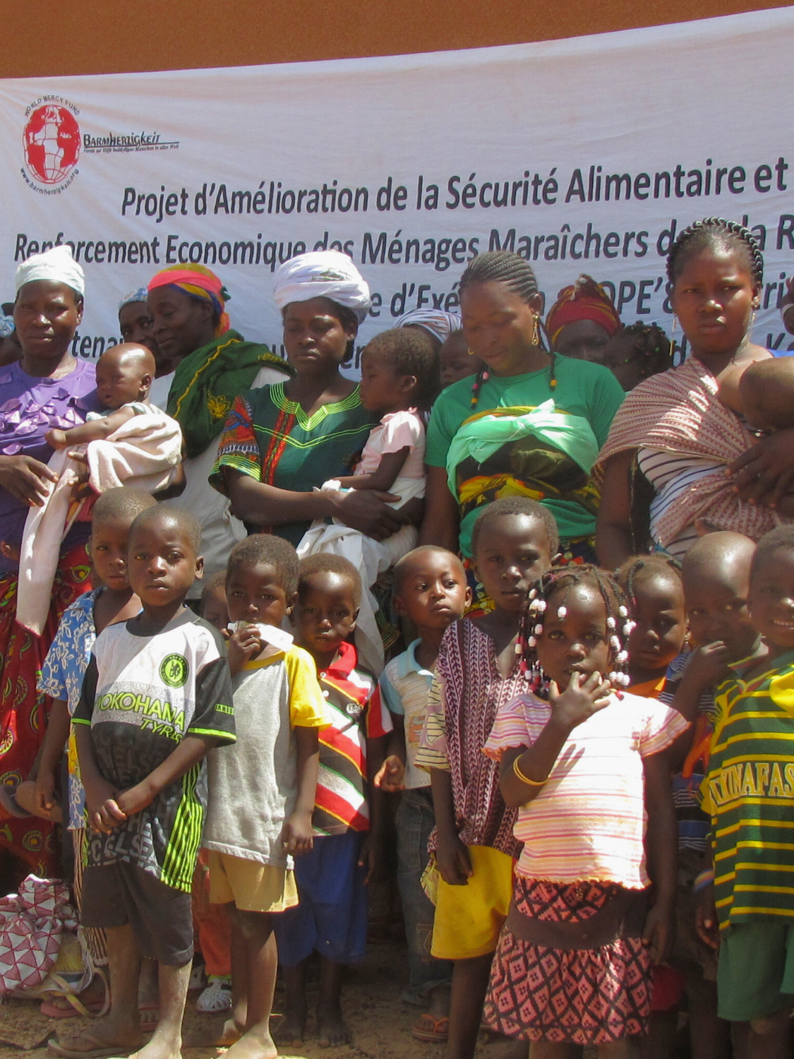Eine große Gruppe von Müttern und Kindern in Burkina Faso steht vor einem Banner eines Ernährungsprojekts. Das Bild zeigt die breite Beteiligung an einem Programm zur Verbesserung der Nahrungssituation für Kinder in Burkina Faso.