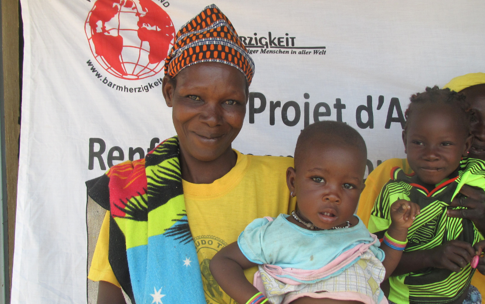 Eine Mutter in Burkina Faso steht mit ihren Kindern vor einem Projektbanner. Das Bild symbolisiert die nachhaltige Hilfe im Bereich „Burkina Faso Nahrung für Kinder“ und zeigt Stolz und Hoffnung durch lokale Unterstützung.