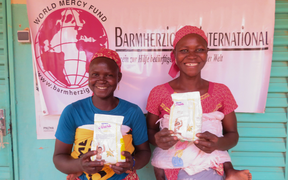 Zwei junge Mütter in Burkina Faso halten lächelnd lokal hergestelltes Kindermehl in der Hand – ein sichtbarer Erfolg des Projekts „Burkina Faso Nahrung für Kinder“ zur Verbesserung der Ernährungssicherheit.