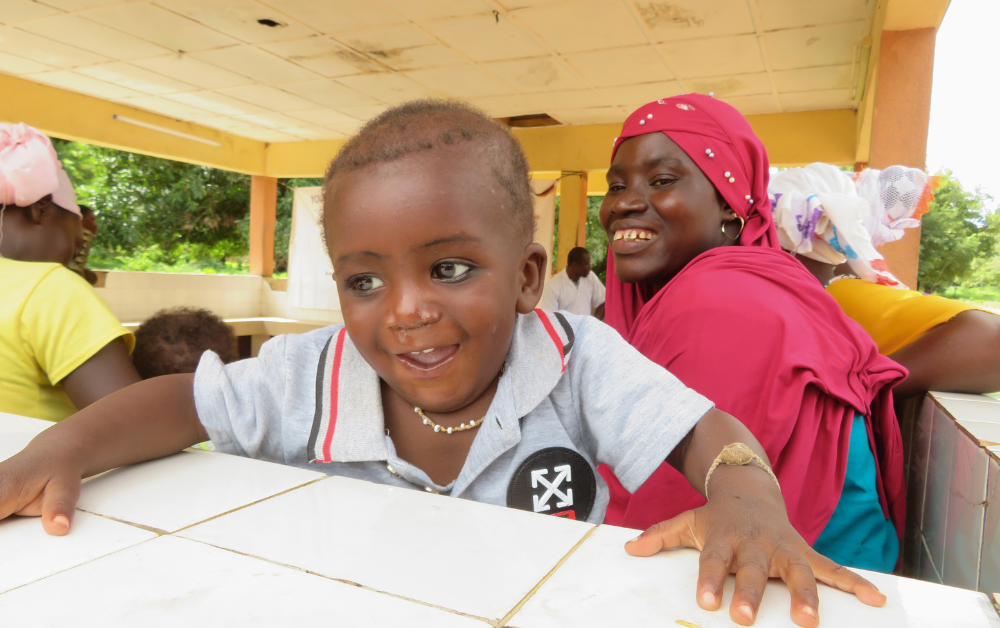 Ein gesund wirkendes Kleinkind aus Burkina Faso lehnt sich lachend über eine Tischkante, im Hintergrund lächelt seine Mutter. Das Projekt „Burkina Faso Nahrung für Kinder“ zeigt Wirkung.