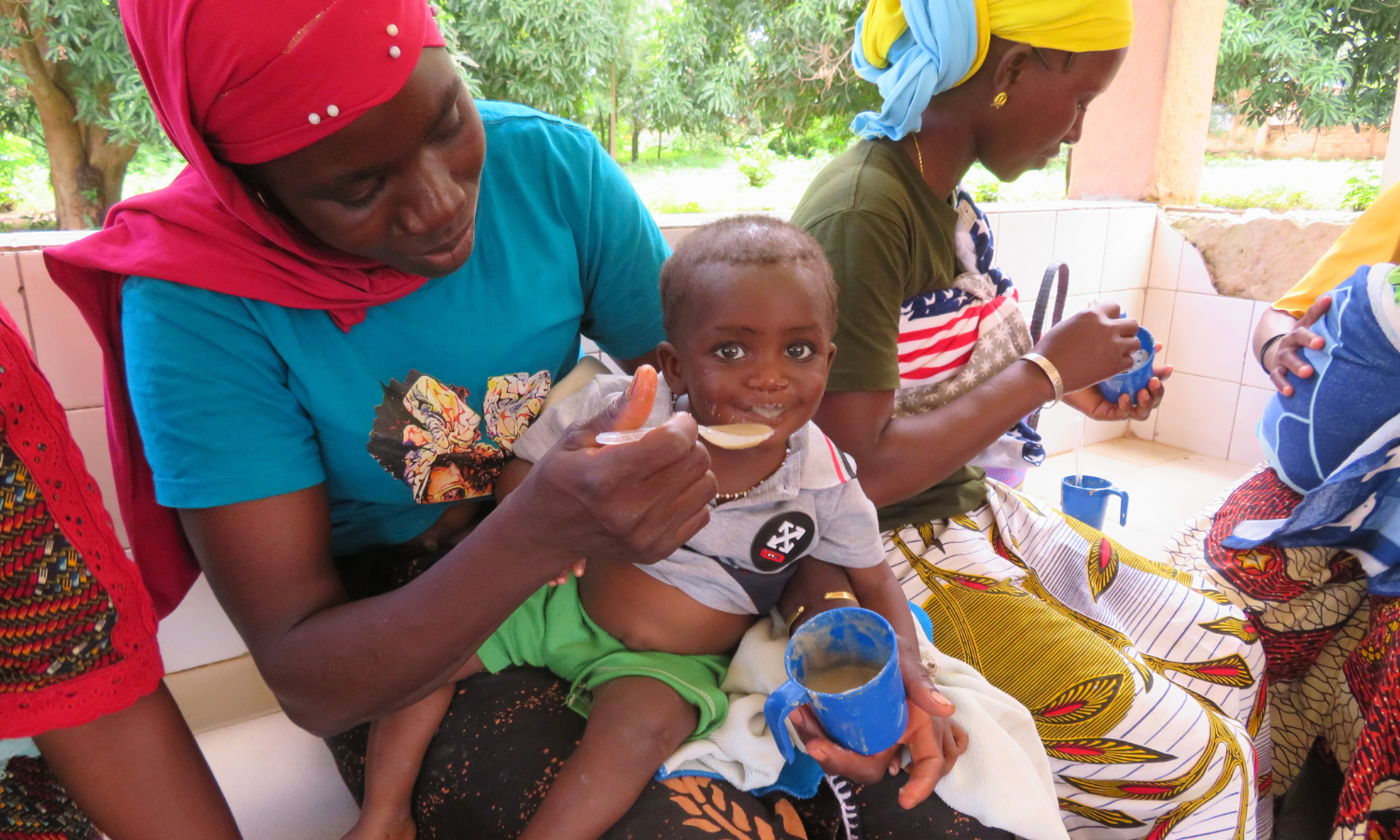 Eine Mutter in Burkina Faso füttert ihr Kleinkind mit nahrhaftem Brei. Das Kind lacht, während es die lokal produzierte Zusatznahrung isst. Ein Beispiel für erfolgreiche Hilfe im Bereich „Burkina Faso Nahrung für Kinder“.