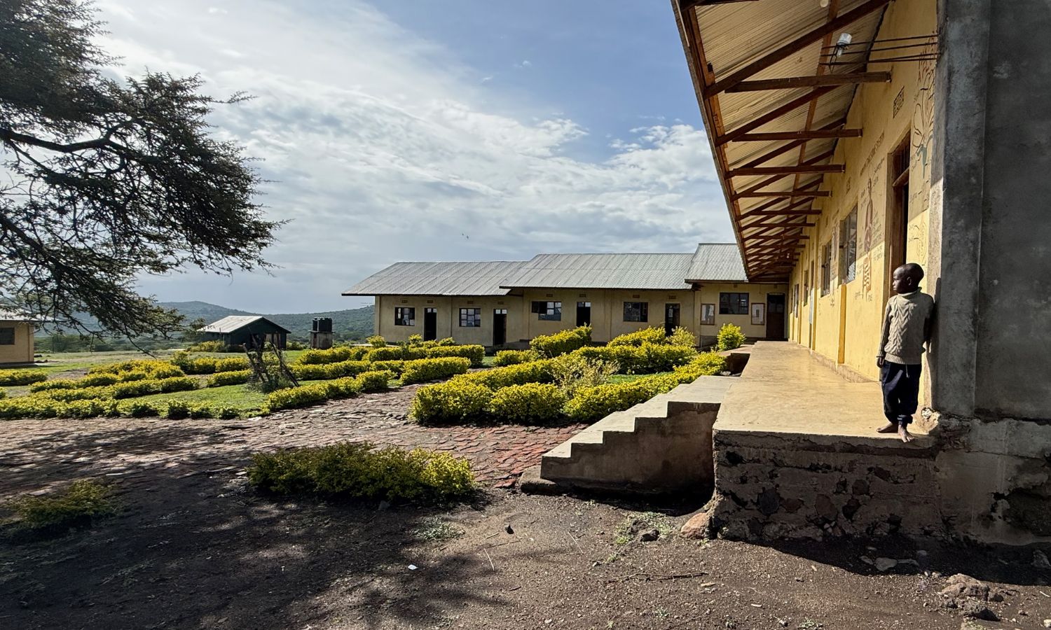 Ein Junge steht vor einem einfachen Schulgebäude der St Luke Pre-Primary School in Endulen, Tansania. Im Hintergrund sind die weite Savannenlandschaft und Hügel nahe der Serengeti zu sehen.