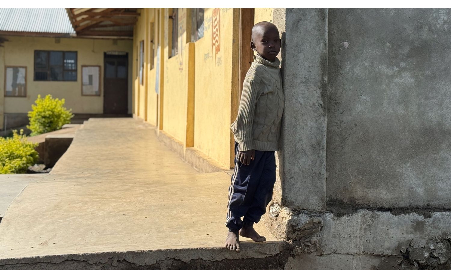 Ein Kind steht barfuß an der Ecke eines Schulgebäudes auf einem erhöhten Gehweg. Im Hintergrund sind weitere Klassenräume mit gelben Wänden und Türen zu sehen.