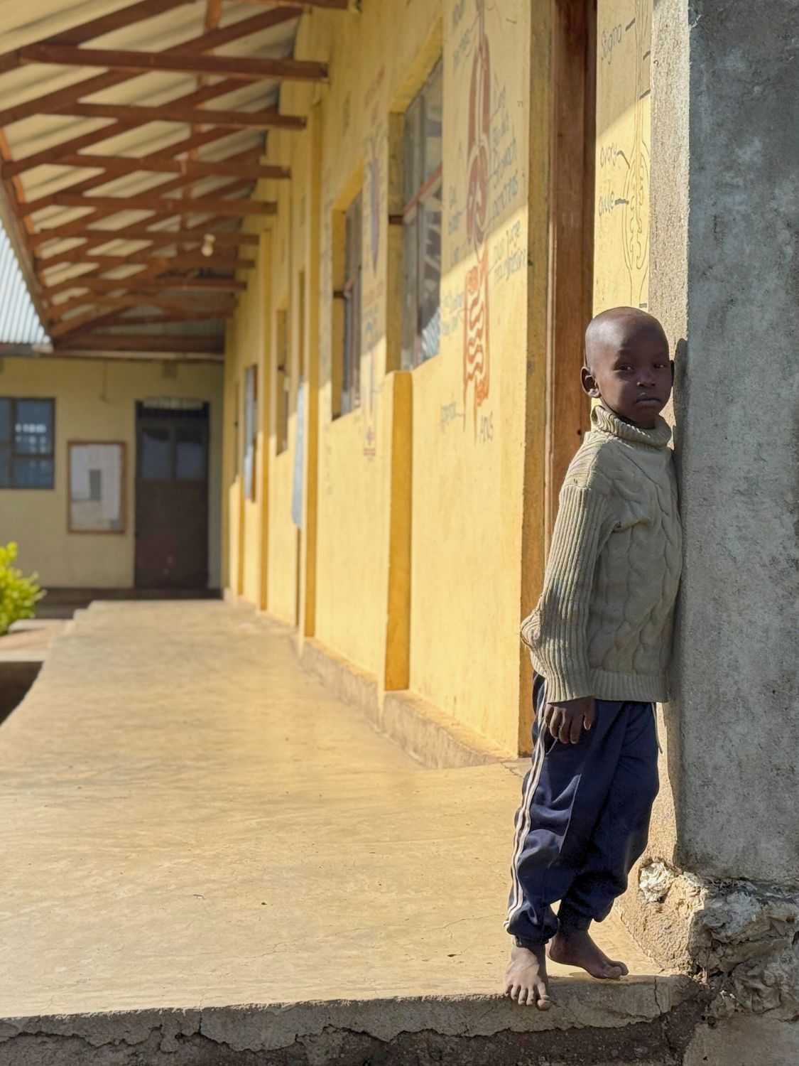 Ein Kind steht barfuß an der Ecke eines Schulgebäudes auf einem erhöhten Gehweg. Im Hintergrund sind weitere Klassenräume mit gelben Wänden und Türen zu sehen.