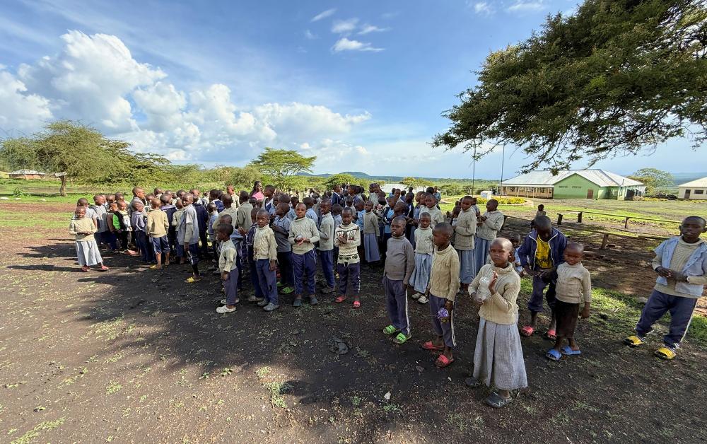 Gruppe von Schulkindern steht in Reihen auf einem offenen Gelände in einer Savannenlandschaft nahe der Serengeti. Im Hintergrund sind flache Schulgebäude, Bäume und weiter Himmel zu sehen.