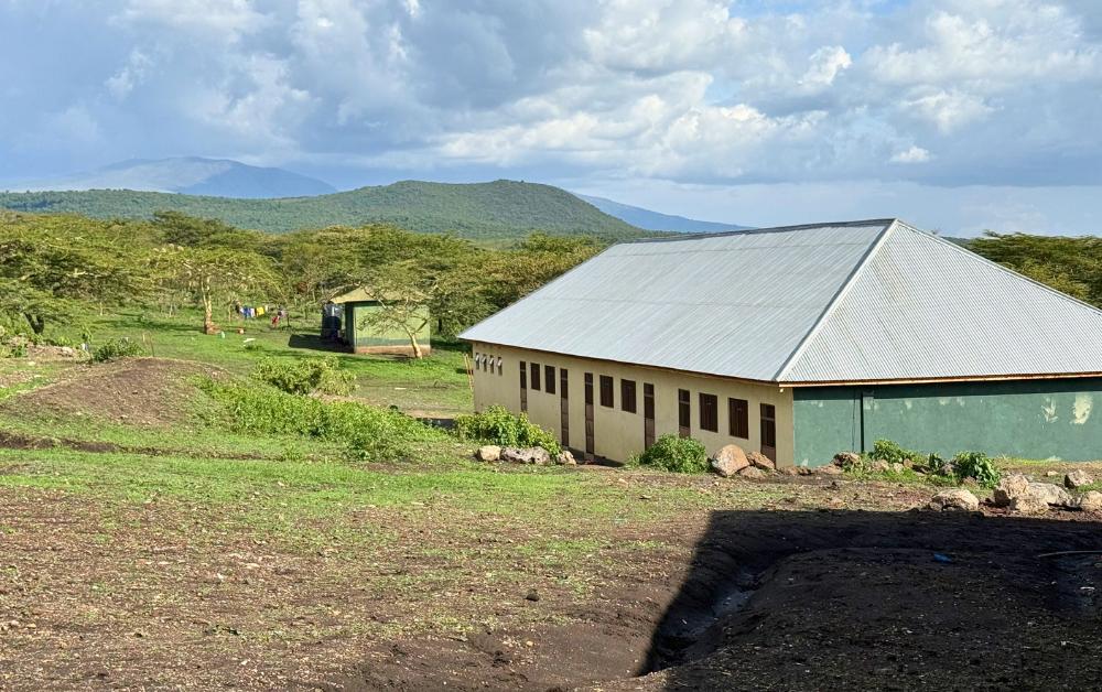 Ein eingeschossiges Schulgebäude mit Wellblechdach steht in einer ländlichen Umgebung. Im Hintergrund sind Bäume, Felder und Hügel zu sehen.