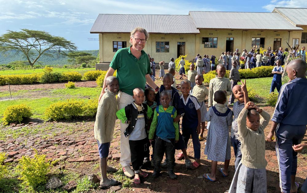 Vorstand Fritz Harms steht auf dem Gelände der St Luke Pre-Primary School in Tansania gemeinsam mit mehreren Schulkindern. Im Hintergrund sind weitere Kinder, ein Schulgebäude und die Savannenlandschaft am Rand der Serengeti zu sehen.