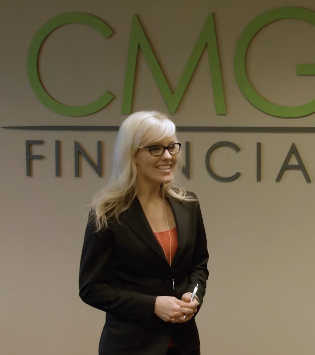 Professional woman standing in front of CMG Financial office wall logo, smiling and holding a pen during a business meeting.