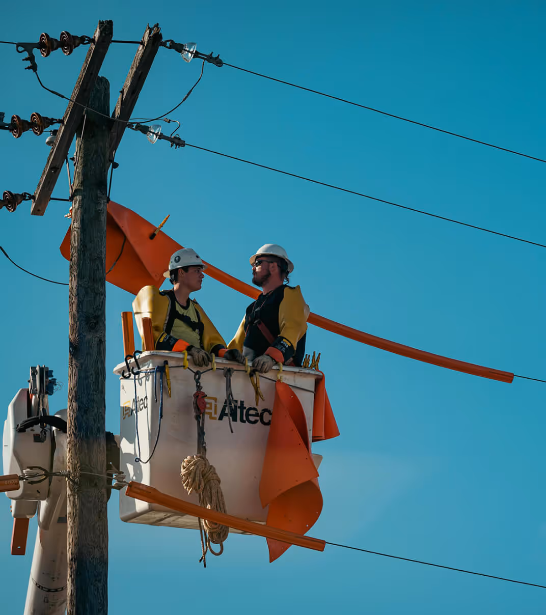 Two utility workers wearing safety gear repairing power lines from an elevated bucket truck under a clear blue sky.
