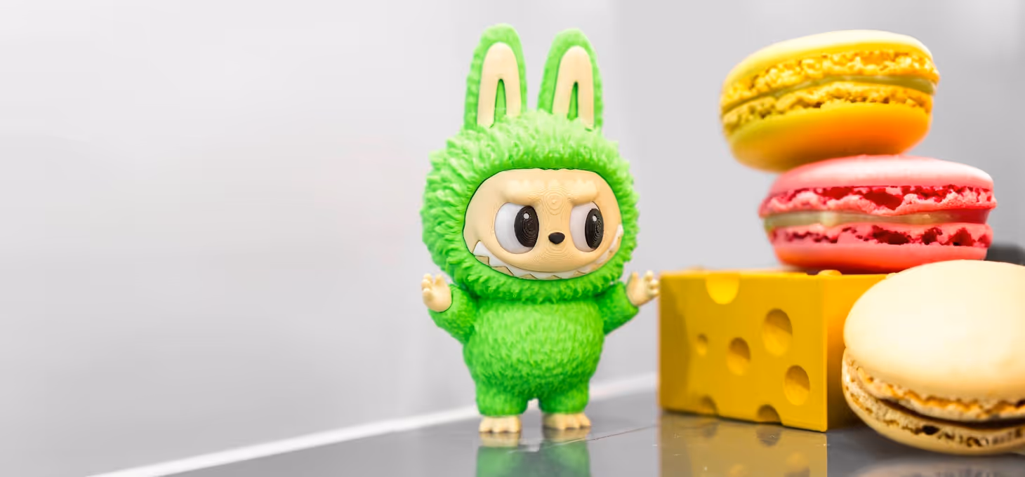 A green Labubu figurine stands beside colourful macarons and a yellow cheese block display.