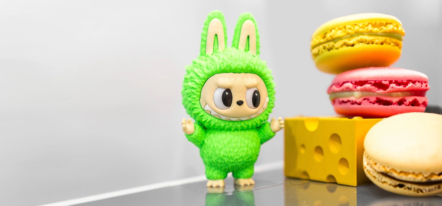 A green Labubu figurine stands beside colourful macarons and a yellow cheese block display.