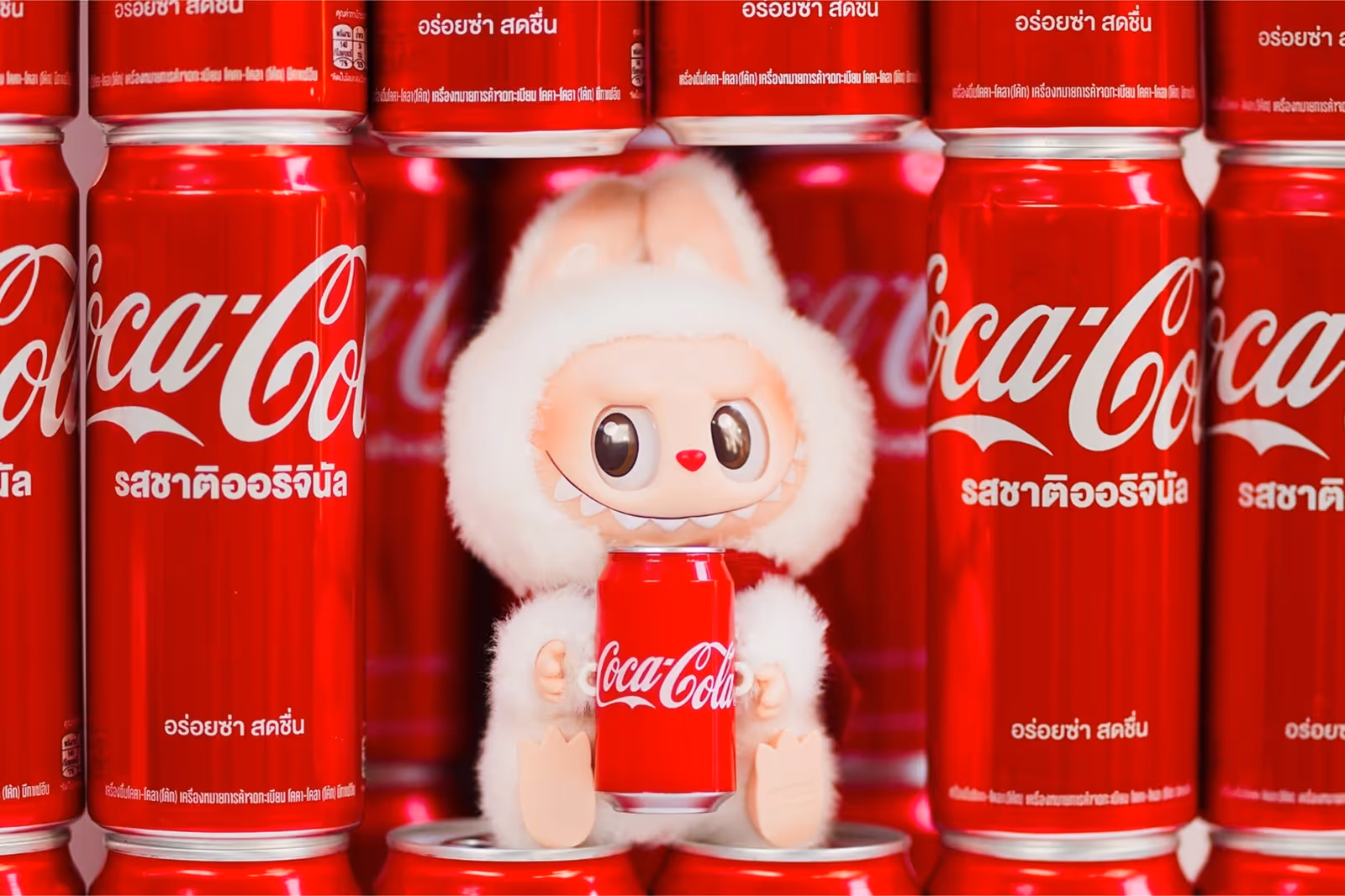 A white Labubu doll holding a Coca-Cola can, surrounded by red Coca-Cola cans, a creative product collaboration between Pop Mart and Coca-Cola.