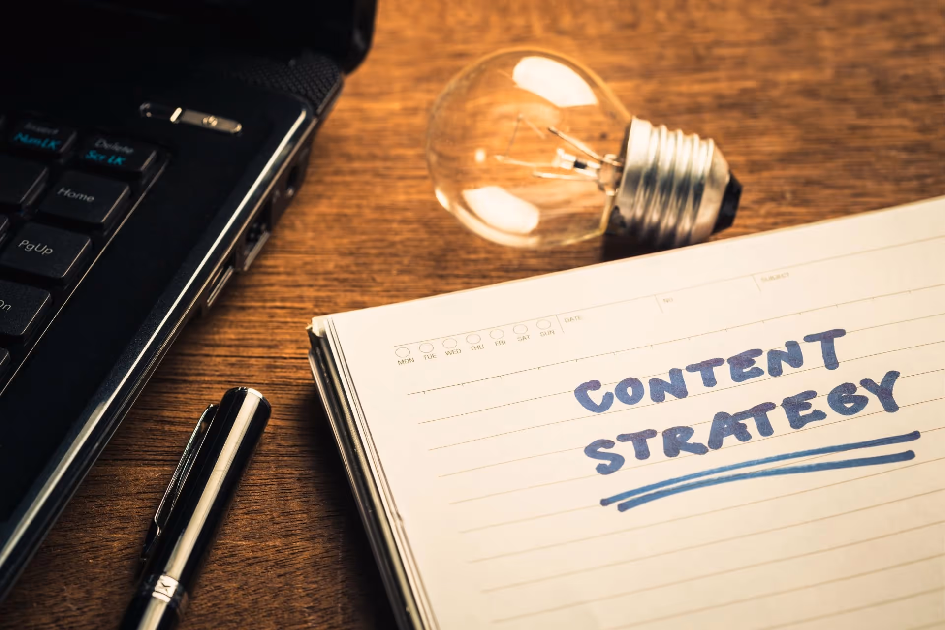 A notebook with “Content Strategy” written on it placed beside a laptop, a lightbulb, and a pen