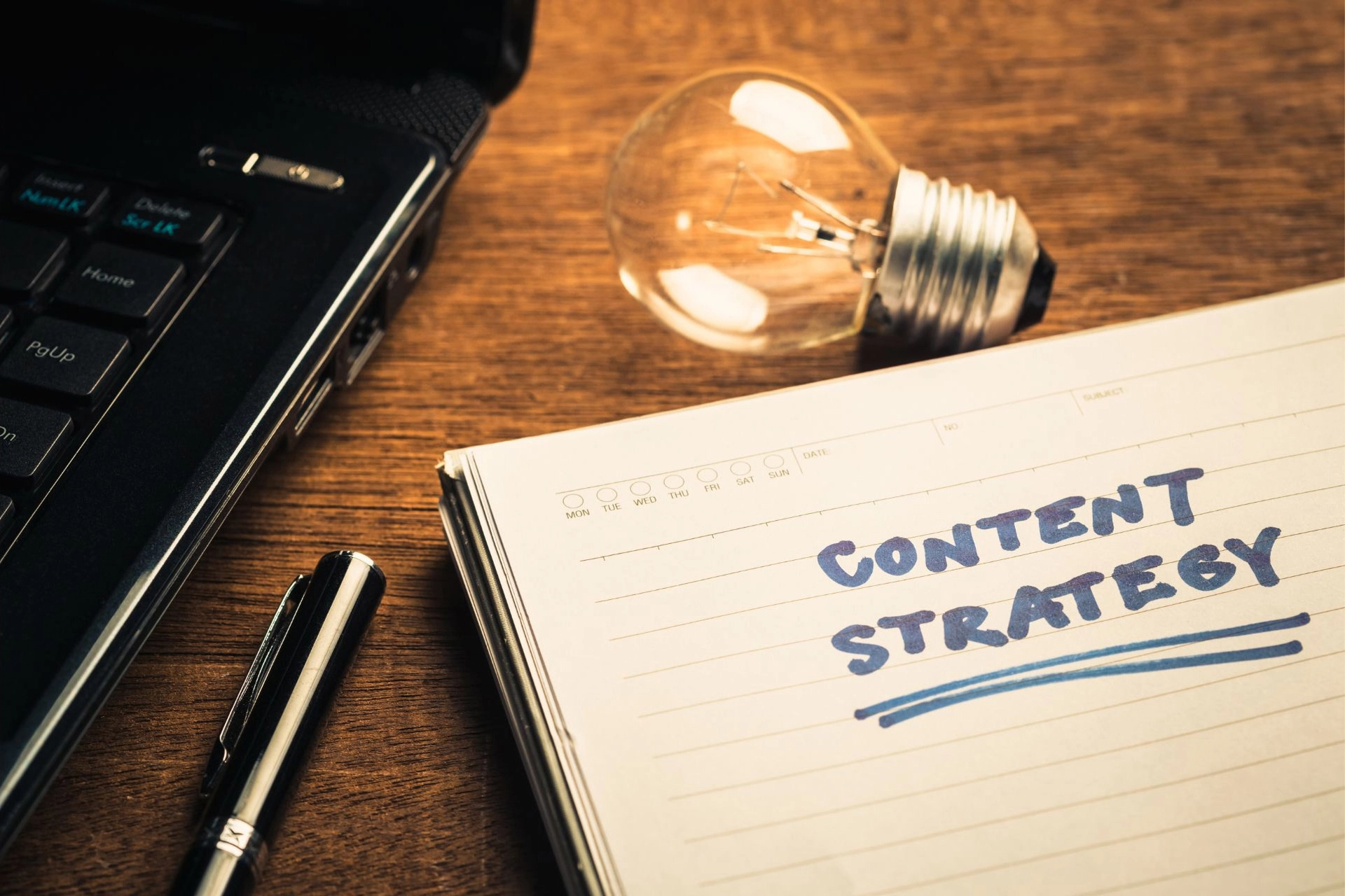 A notebook with “Content Strategy” written on it placed beside a laptop, a lightbulb, and a pen