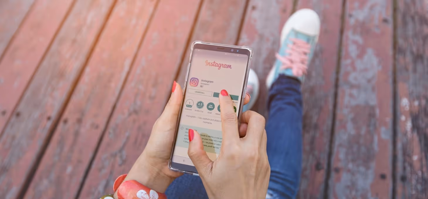 A woman’s lower body is shown sitting on a wooden deck holding a phone showing Instagram.