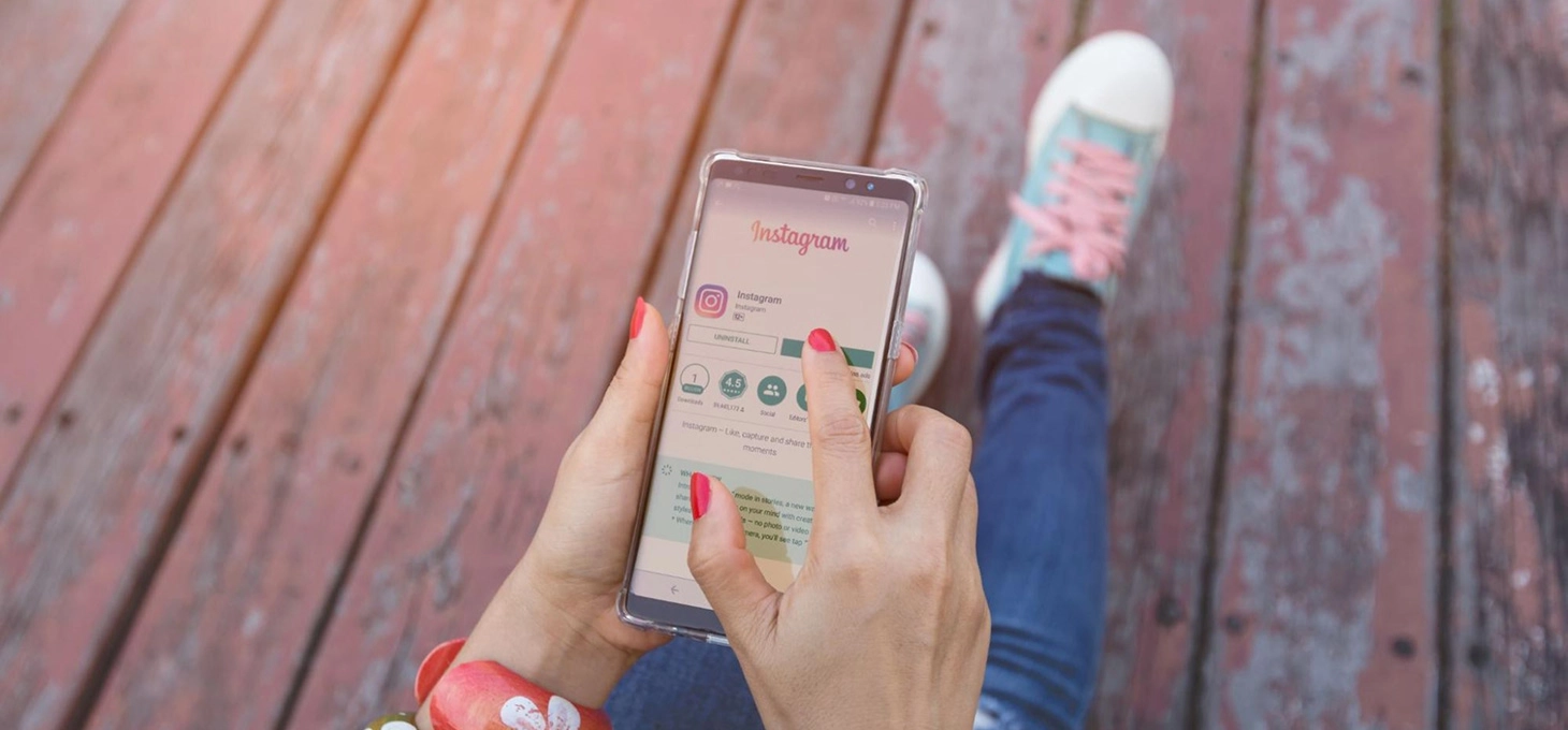 A woman’s lower body is shown sitting on a wooden deck holding a phone showing Instagram.