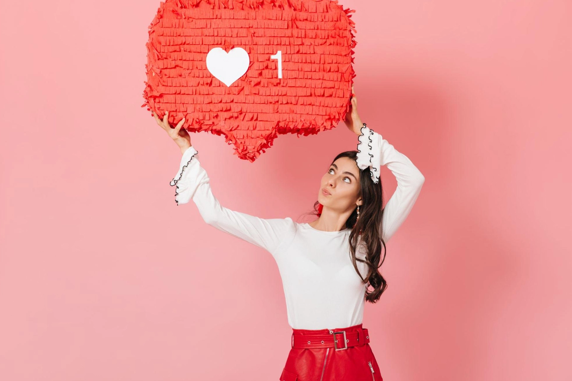 A young woman stands against a pink background, holding up a papier-mâché Instagram comment bubble.