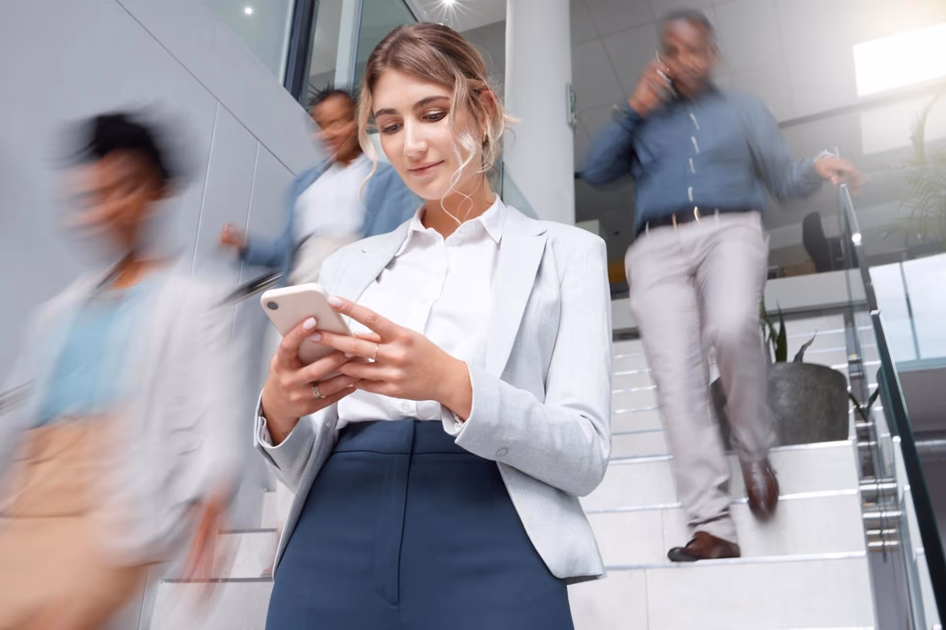A woman in a white blazer looks at her phone as she descends a staircase.