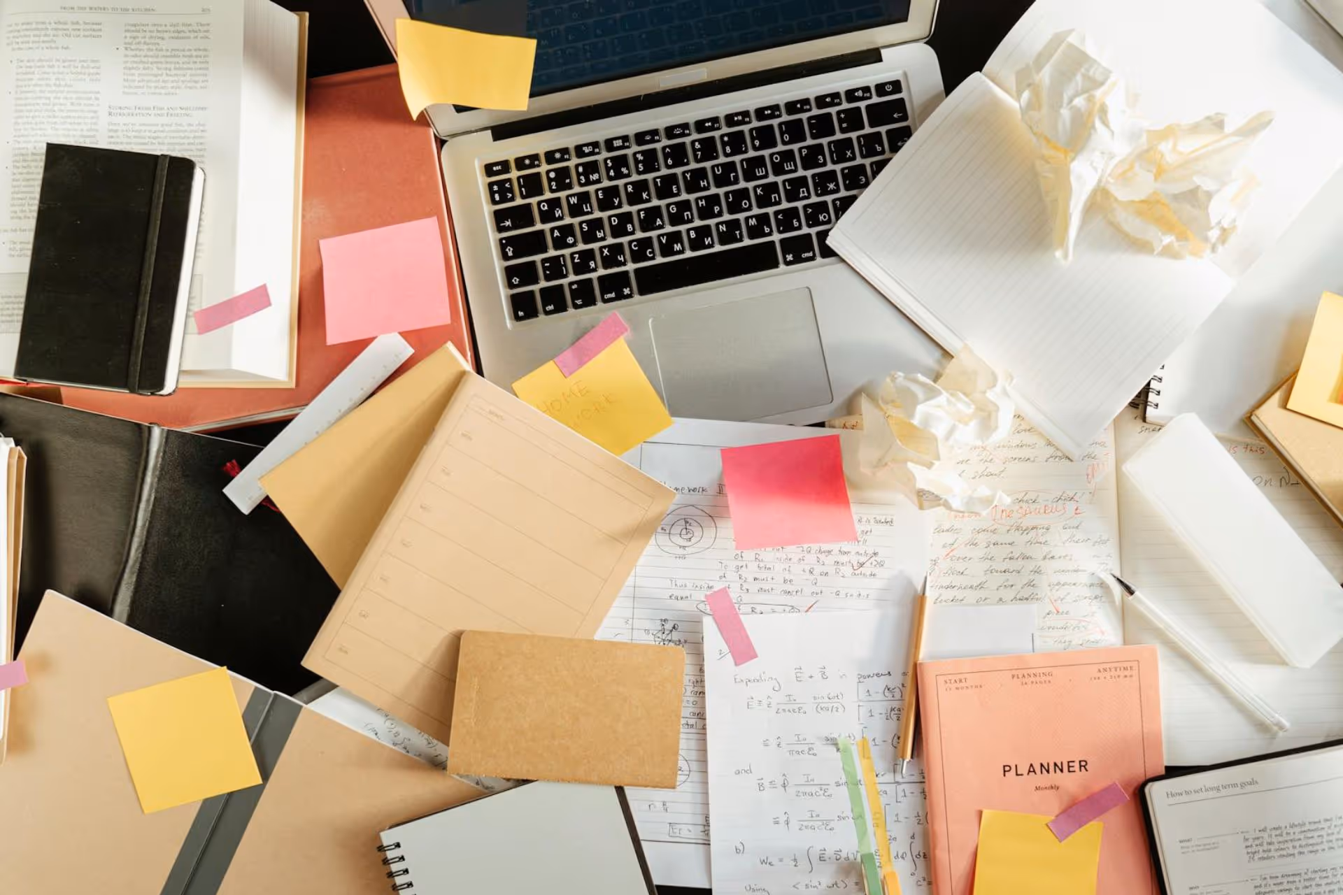 A messy desk with several notebooks and notepads beside a Mac