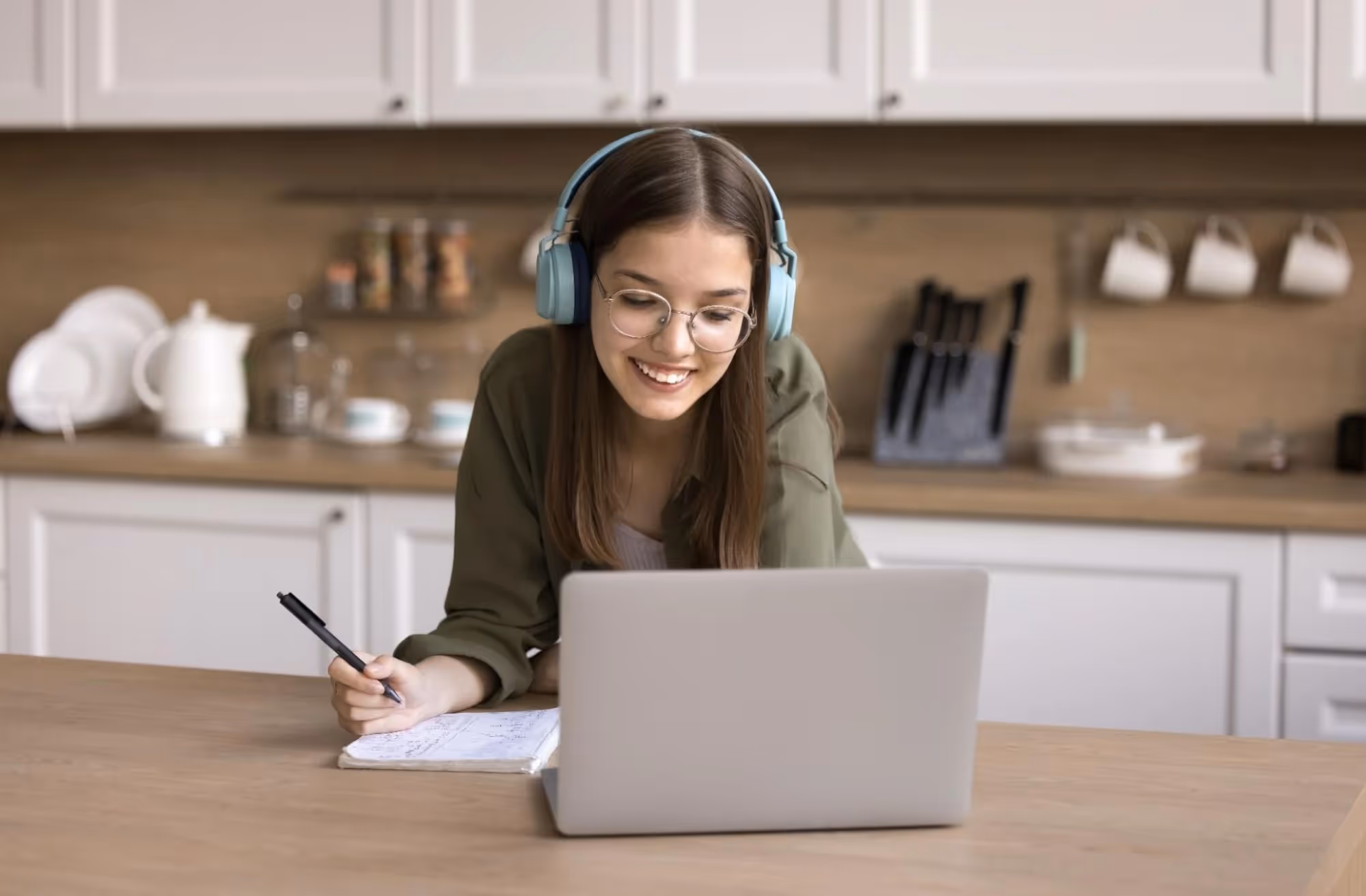 A young woman in a kitchen smiles at her laptop, headphones on.