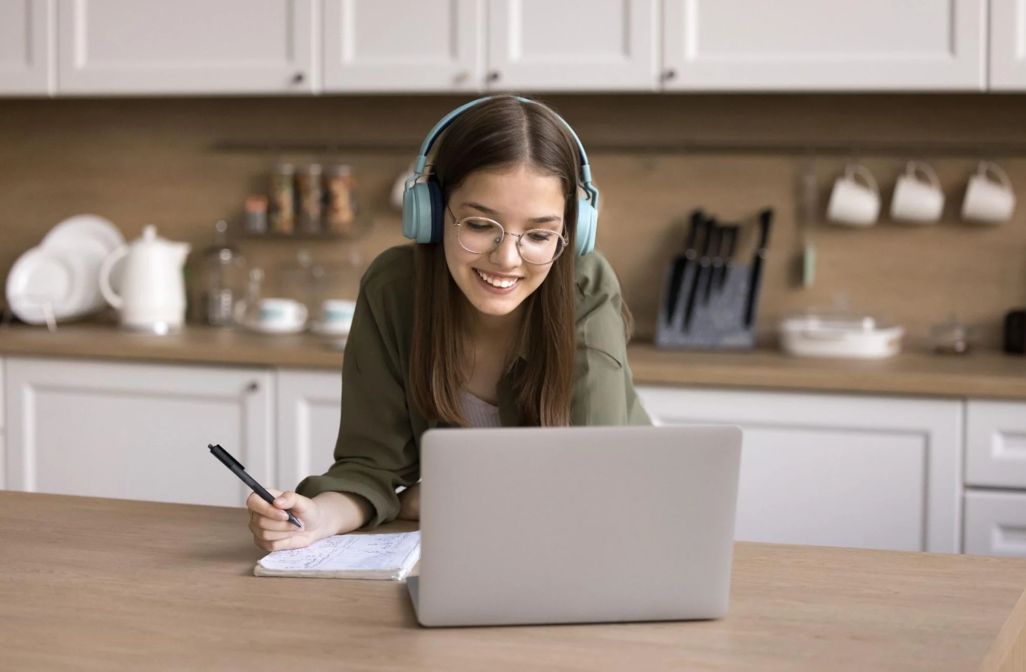 A young woman in a kitchen smiles at her laptop, headphones on.