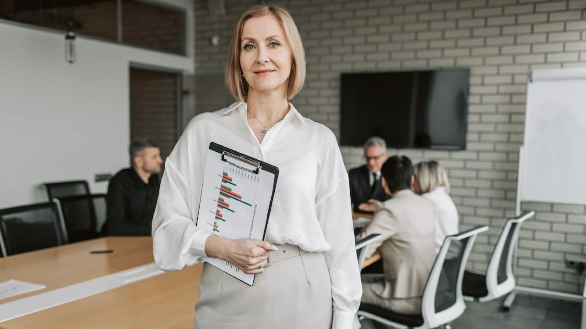 A professional woman stands in a conference room holding a clipboard, with a meeting taking place in the background