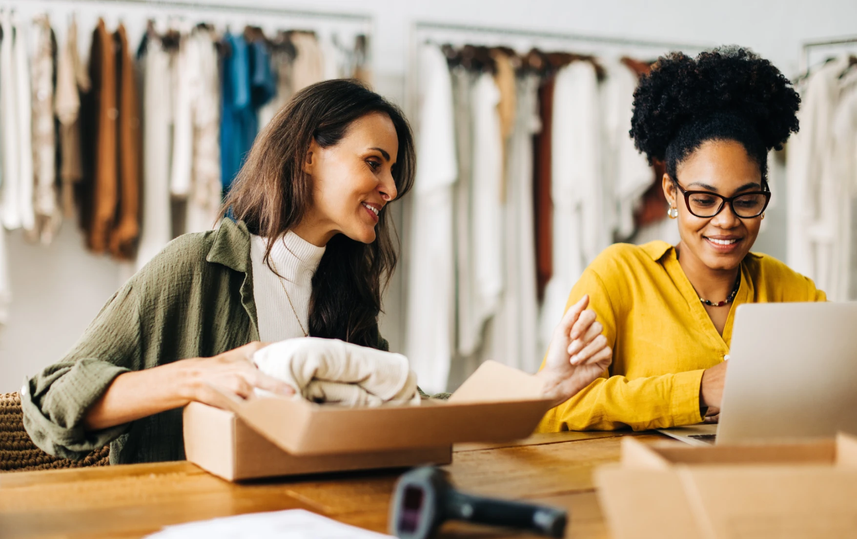A businesswoman checking ecommerce website on a laptop beside another woman packing clothes in a delivery box