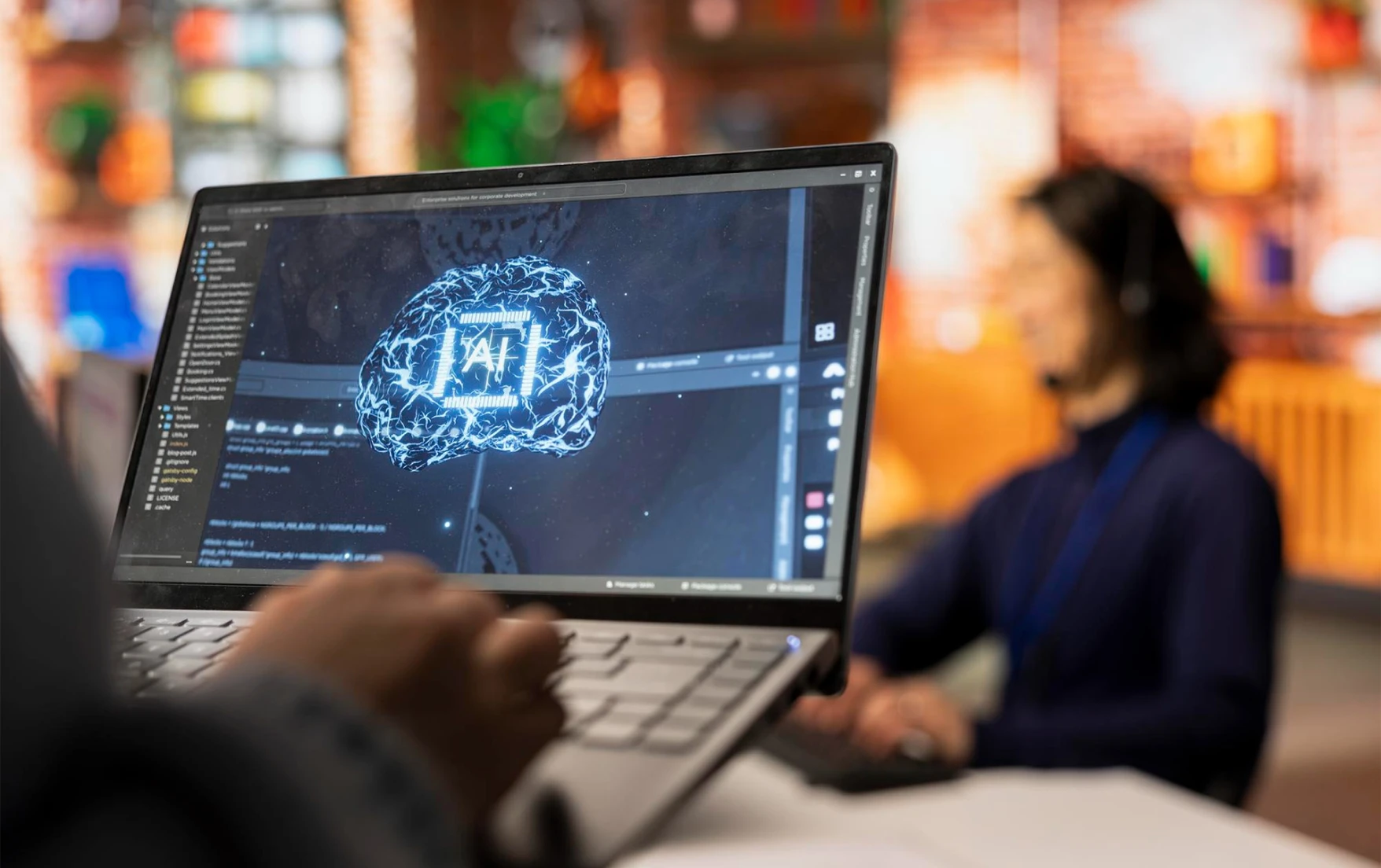 a person uses a laptop displaying a digital brain with “AI” text. A blurred coworker in the background.