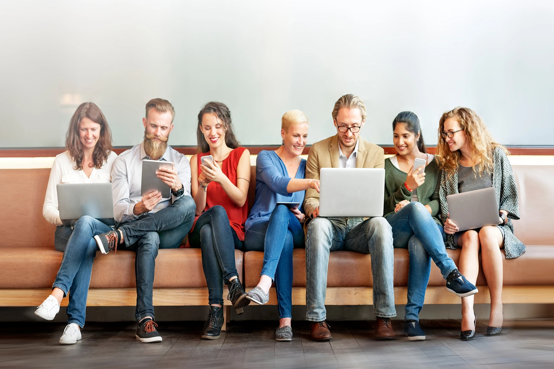 people of diverse ages and backgrounds sitting on a sofa, using assorted digital devices.