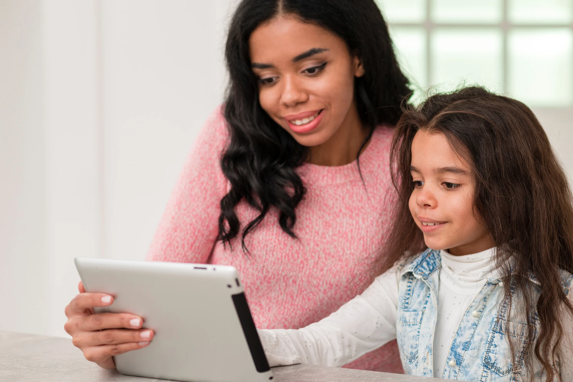 A mother and a child sitting together at a table, focused on a digital tablet. Both look happy and engaged with the content on the screen.