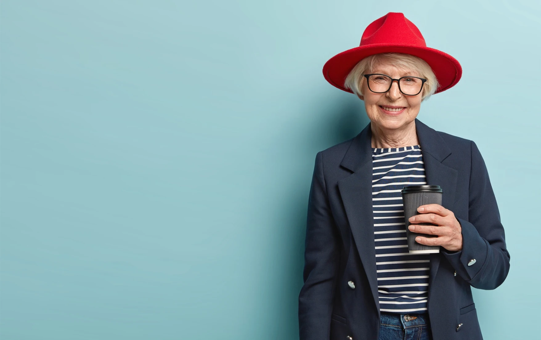 Smiling older woman in glasses and a red hat, wearing a navy blazer and a striped shirt, holds a cup of coffee against a light blue background.